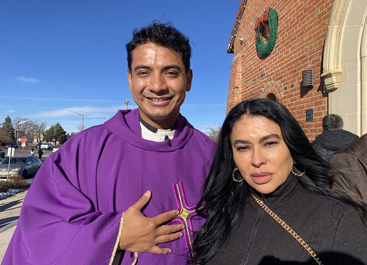 A priest and a woman are standing together, posing for a picture. The man is wearing a purple robe, and the woman is wearing a black shirt. They are both smiling.