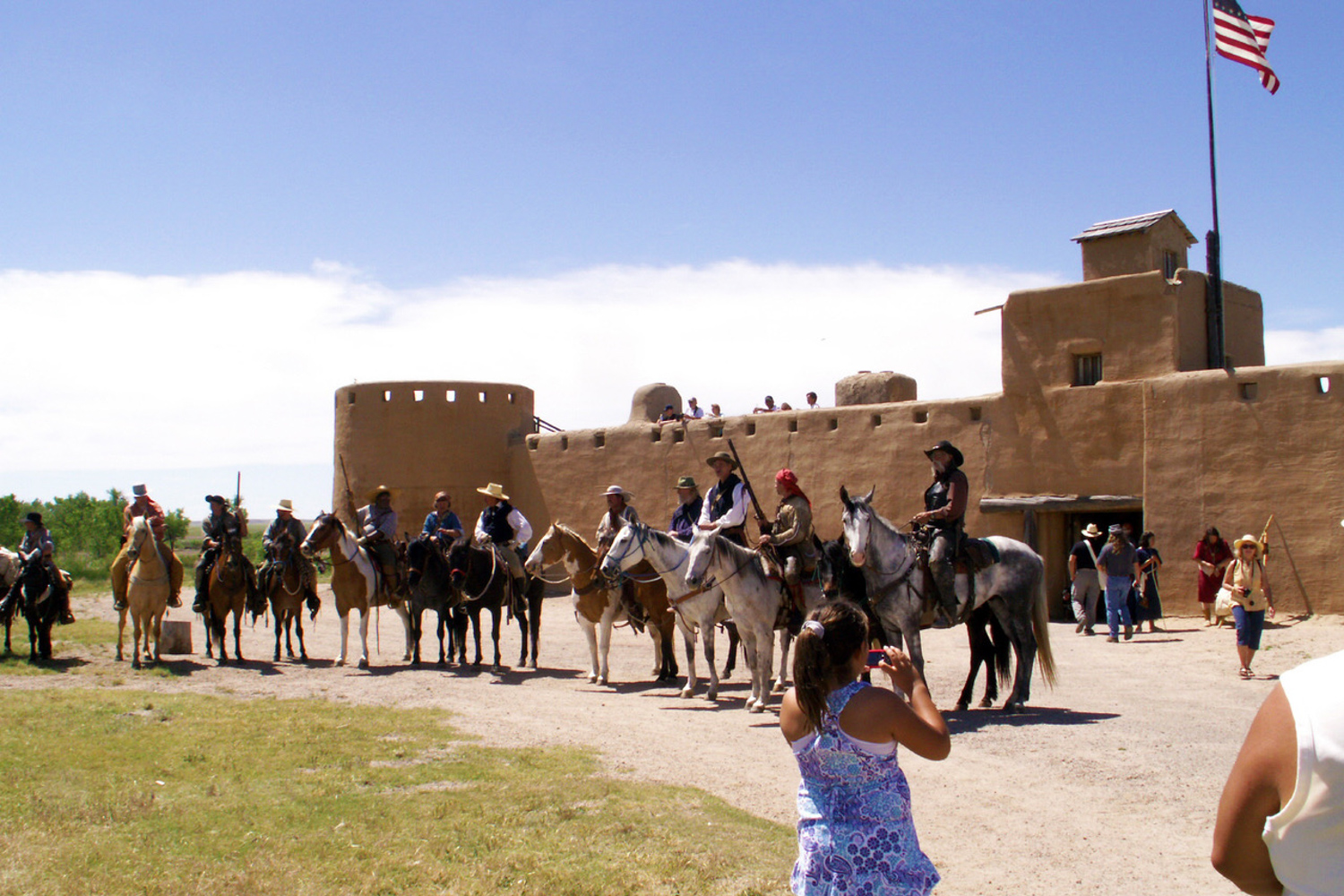 about a dozen people on horseback lined up in front of an adobe building