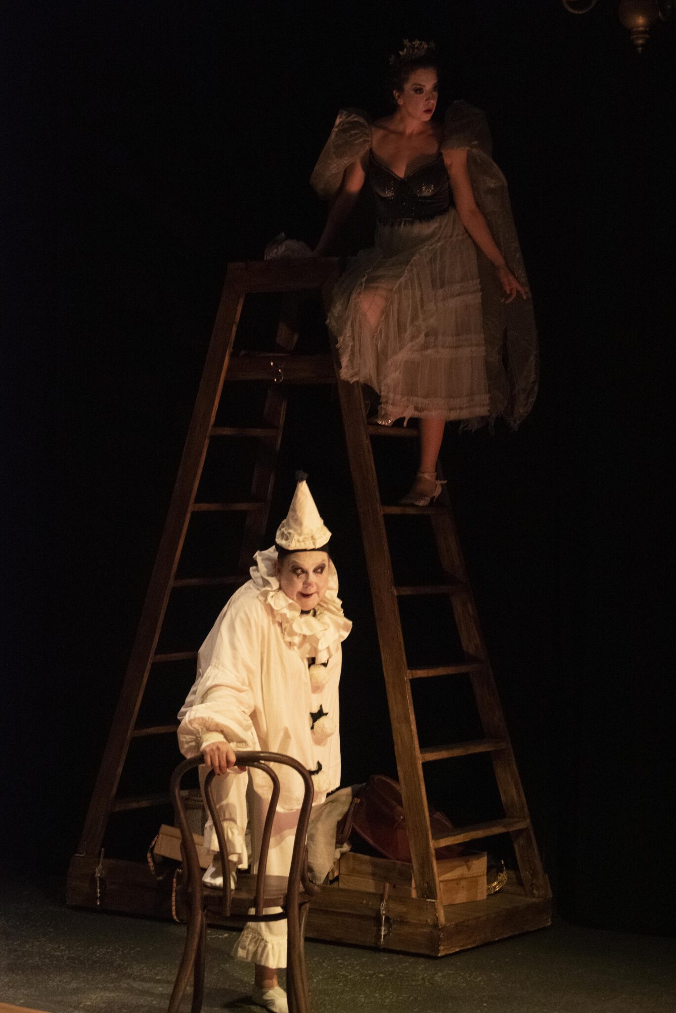 A woman in a clown costume stands menacingly. Above her, an angelic figure floats at the top of a ladder.